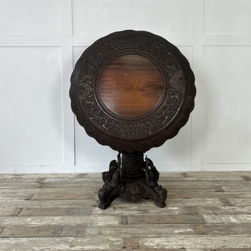 A nineteenth-century dark carved hardwood Burmese tilt-top table shown with the circular top tilted vertically, featuring intricate relief carvings along the border and a heavily sculpted pedestal base.