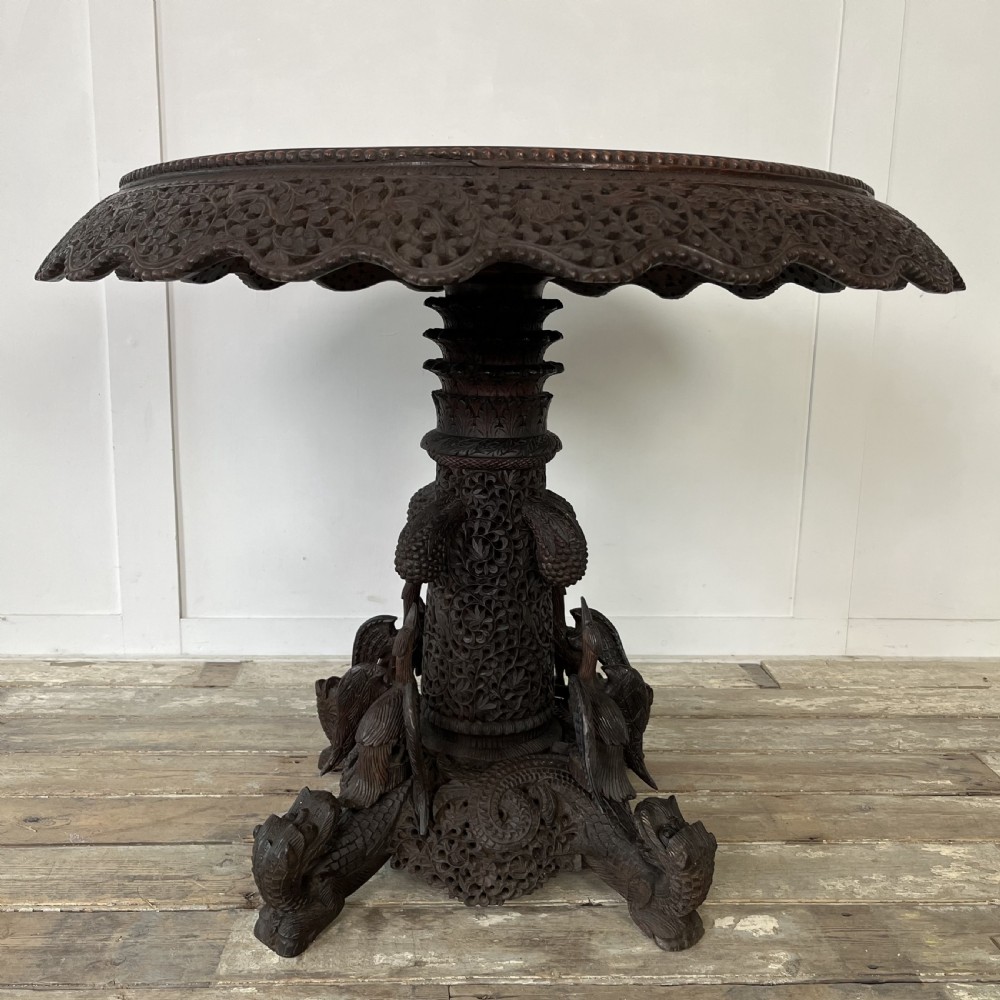 A nineteenth-century dark carved hardwood Burmese tilt-top table shown with the circular top tilted vertically, featuring intricate relief carvings along the border and a heavily sculpted pedestal base. View V