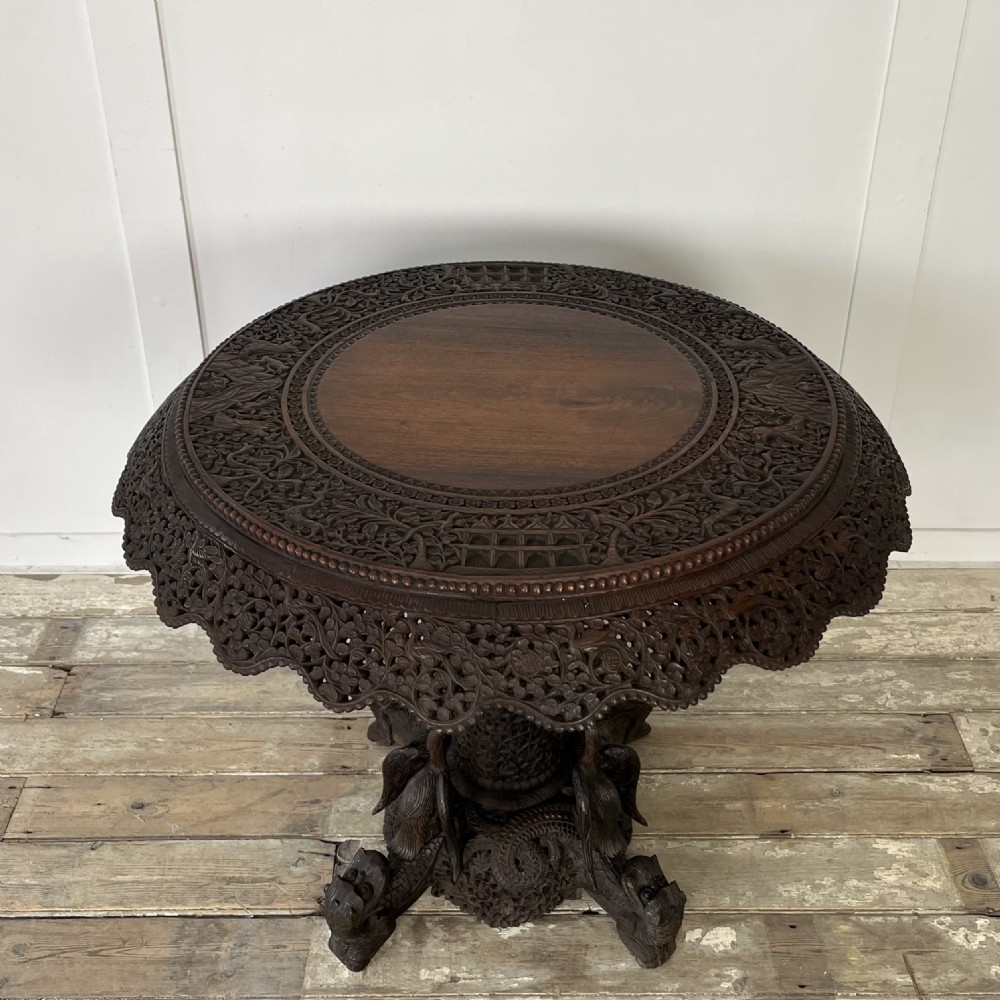 A nineteenth-century dark carved hardwood Burmese tilt-top table shown with the circular top tilted vertically, featuring intricate relief carvings along the border and a heavily sculpted pedestal base. View VI