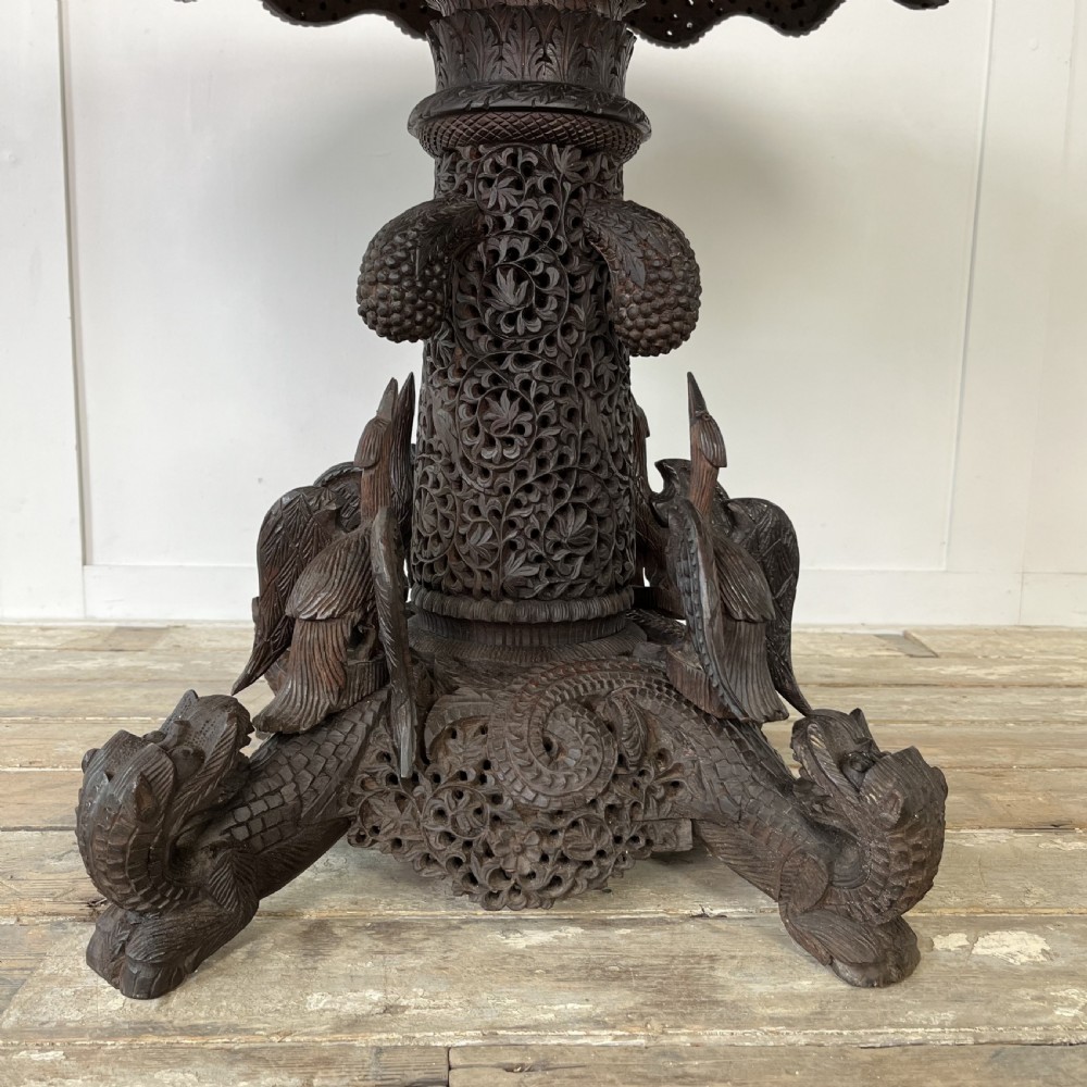 A nineteenth-century dark carved hardwood Burmese tilt-top table shown with the circular top tilted vertically, featuring intricate relief carvings along the border and a heavily sculpted pedestal base. View VIII