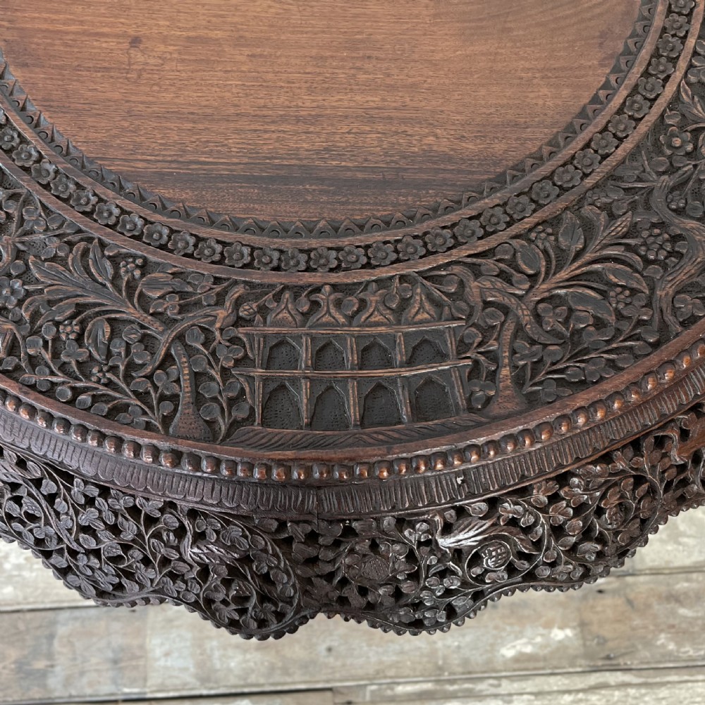 A nineteenth-century dark carved hardwood Burmese tilt-top table shown with the circular top tilted vertically, featuring intricate relief carvings along the border and a heavily sculpted pedestal base. View XIII