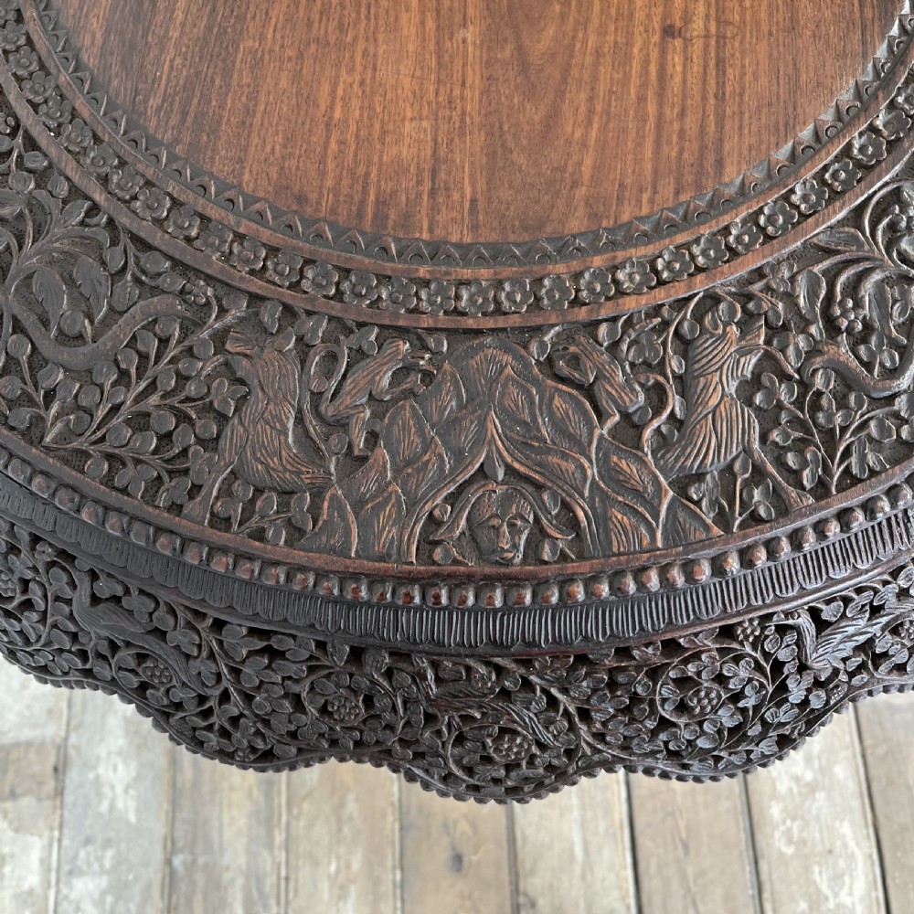 A nineteenth-century dark carved hardwood Burmese tilt-top table shown with the circular top tilted vertically, featuring intricate relief carvings along the border and a heavily sculpted pedestal base. View XIV