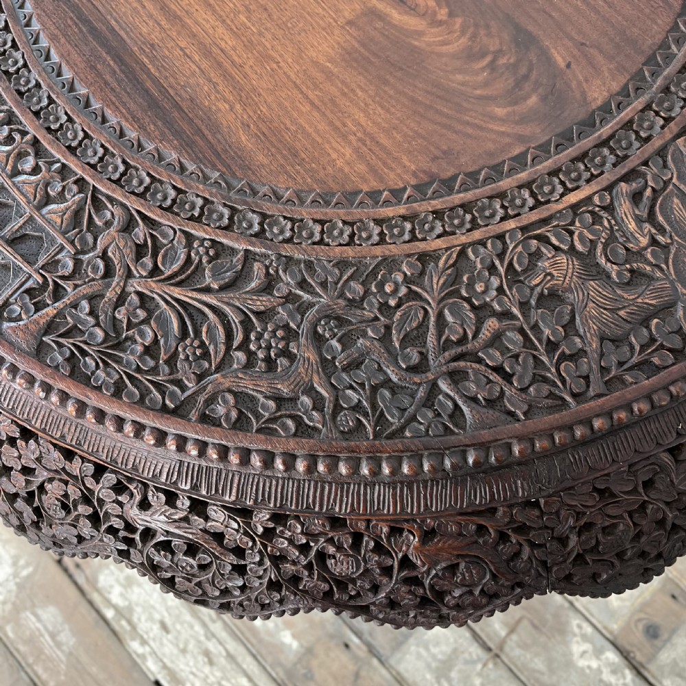 A nineteenth-century dark carved hardwood Burmese tilt-top table shown with the circular top tilted vertically, featuring intricate relief carvings along the border and a heavily sculpted pedestal base. View XV