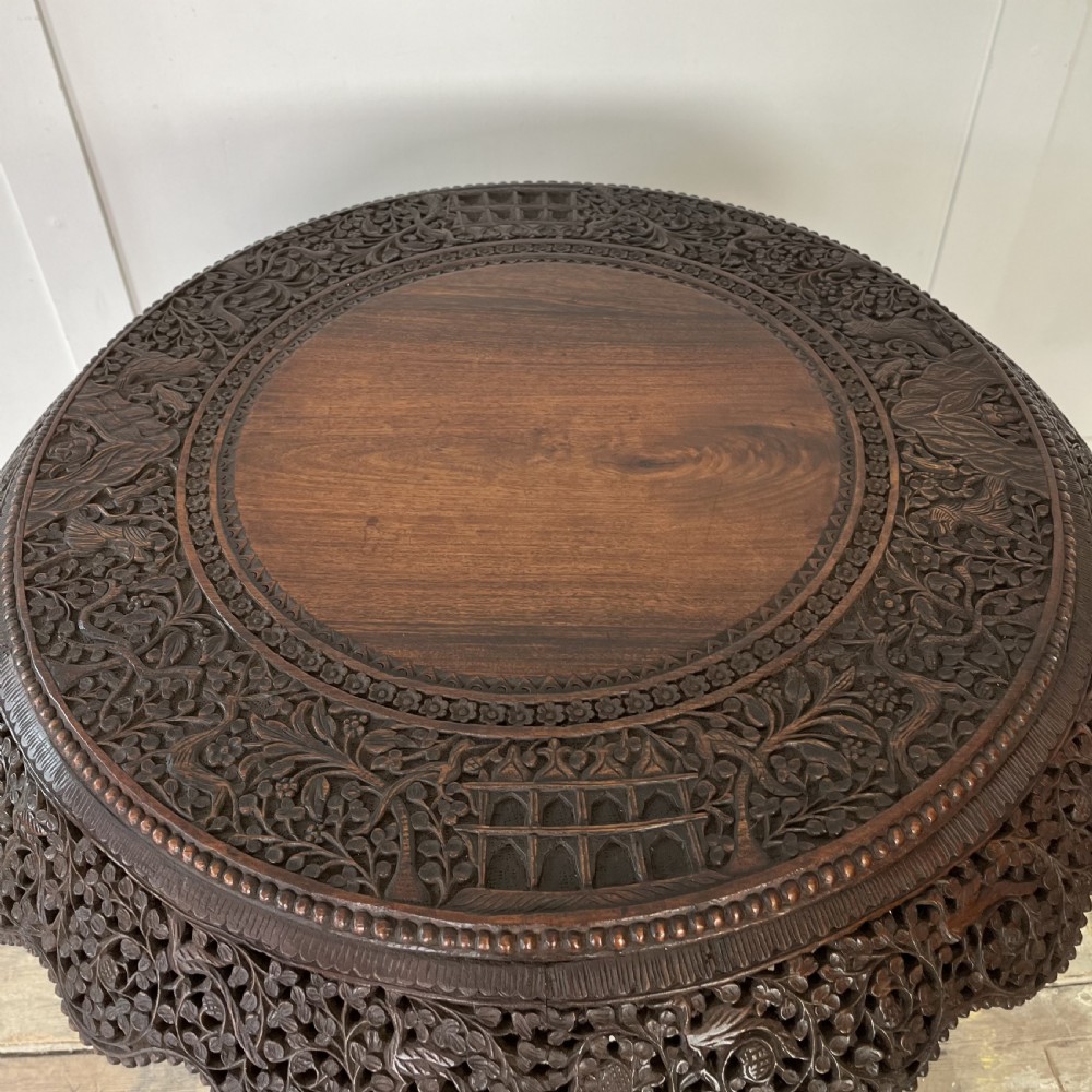 A nineteenth-century dark carved hardwood Burmese tilt-top table shown with the circular top tilted vertically, featuring intricate relief carvings along the border and a heavily sculpted pedestal base. View XVI