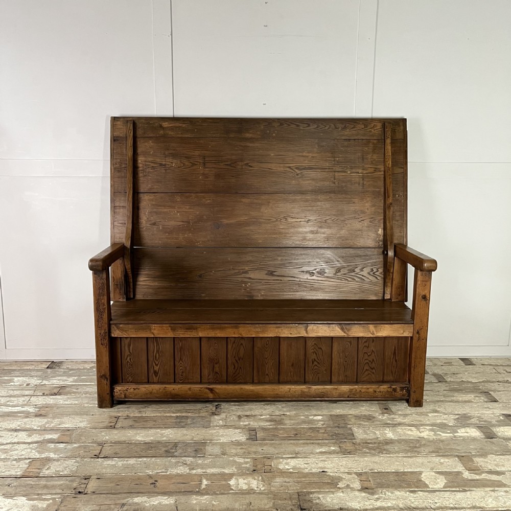A dark stained Victorian pine monks bench featuring a tall planked backrest, flat wooden armrests, and a vertical panelled base.