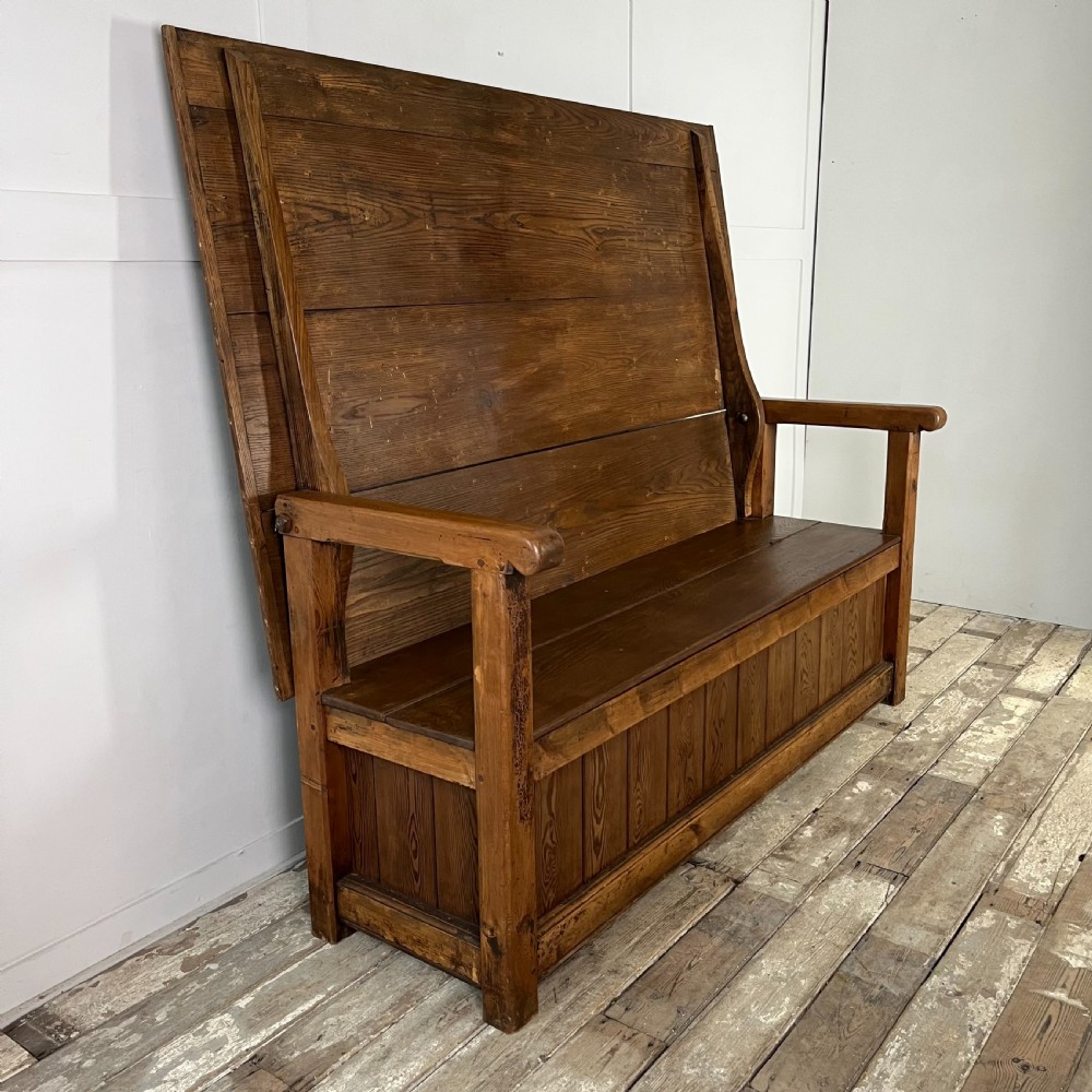 A dark stained Victorian pine monks bench featuring a tall planked backrest, flat wooden armrests, and a vertical panelled base. View VI