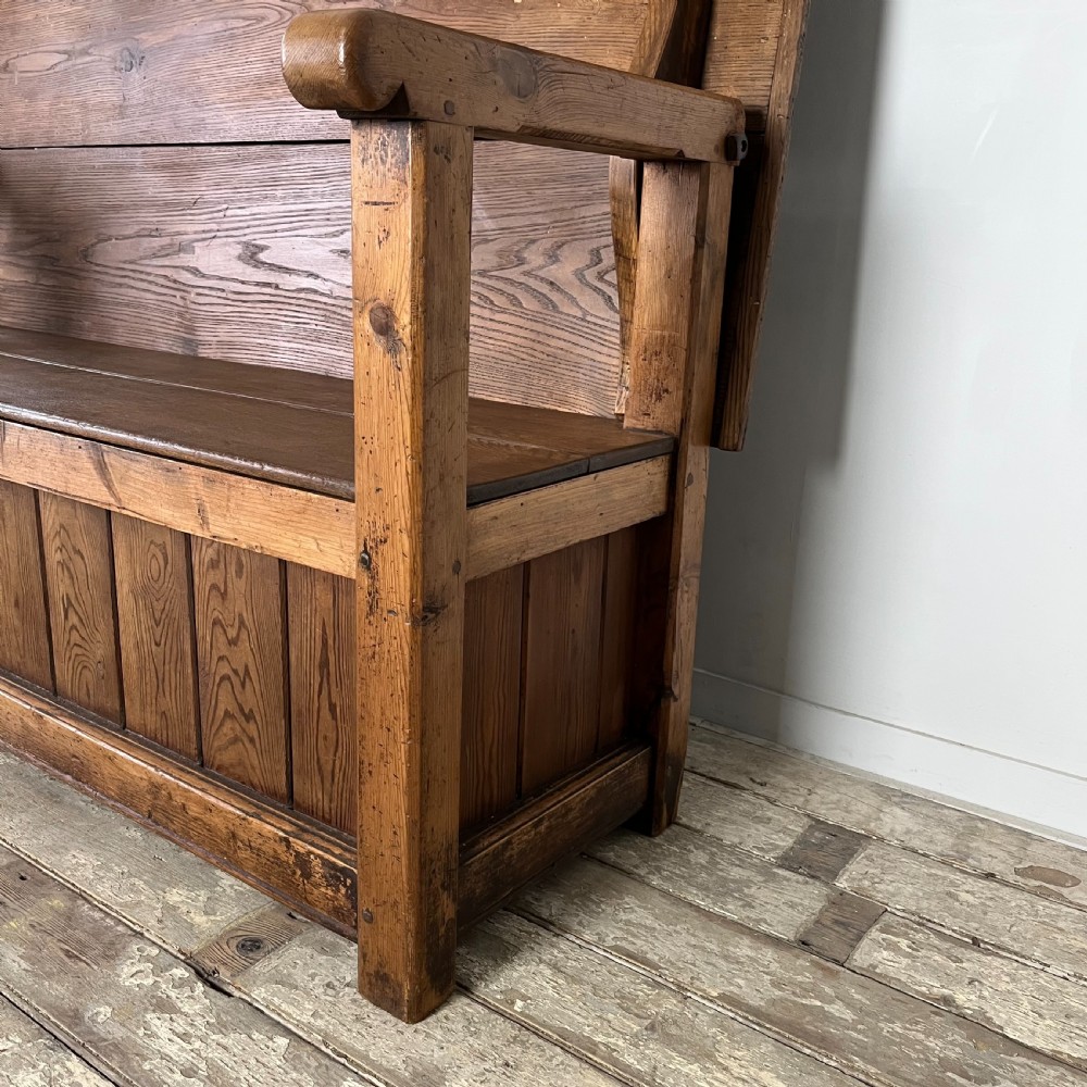 A dark stained Victorian pine monks bench featuring a tall planked backrest, flat wooden armrests, and a vertical panelled base. View VII