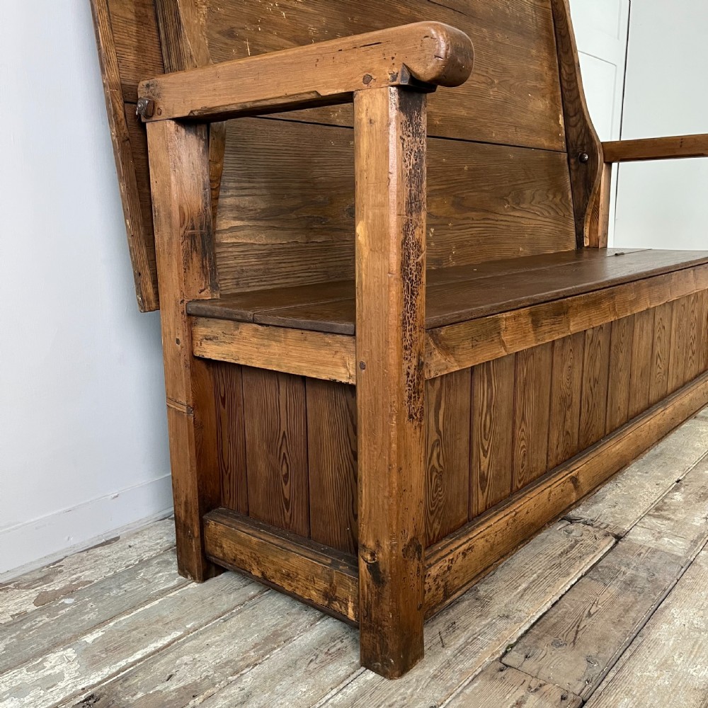 A dark stained Victorian pine monks bench featuring a tall planked backrest, flat wooden armrests, and a vertical panelled base. View IX