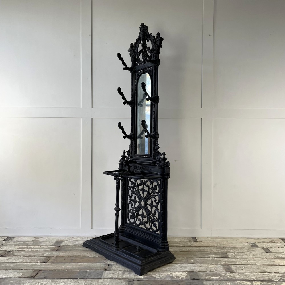 A tall black cast iron Victorian hall stand featuring an integrated central mirror, branching coat hooks, scrollwork tracery, and an umbrella drip tray at the base.