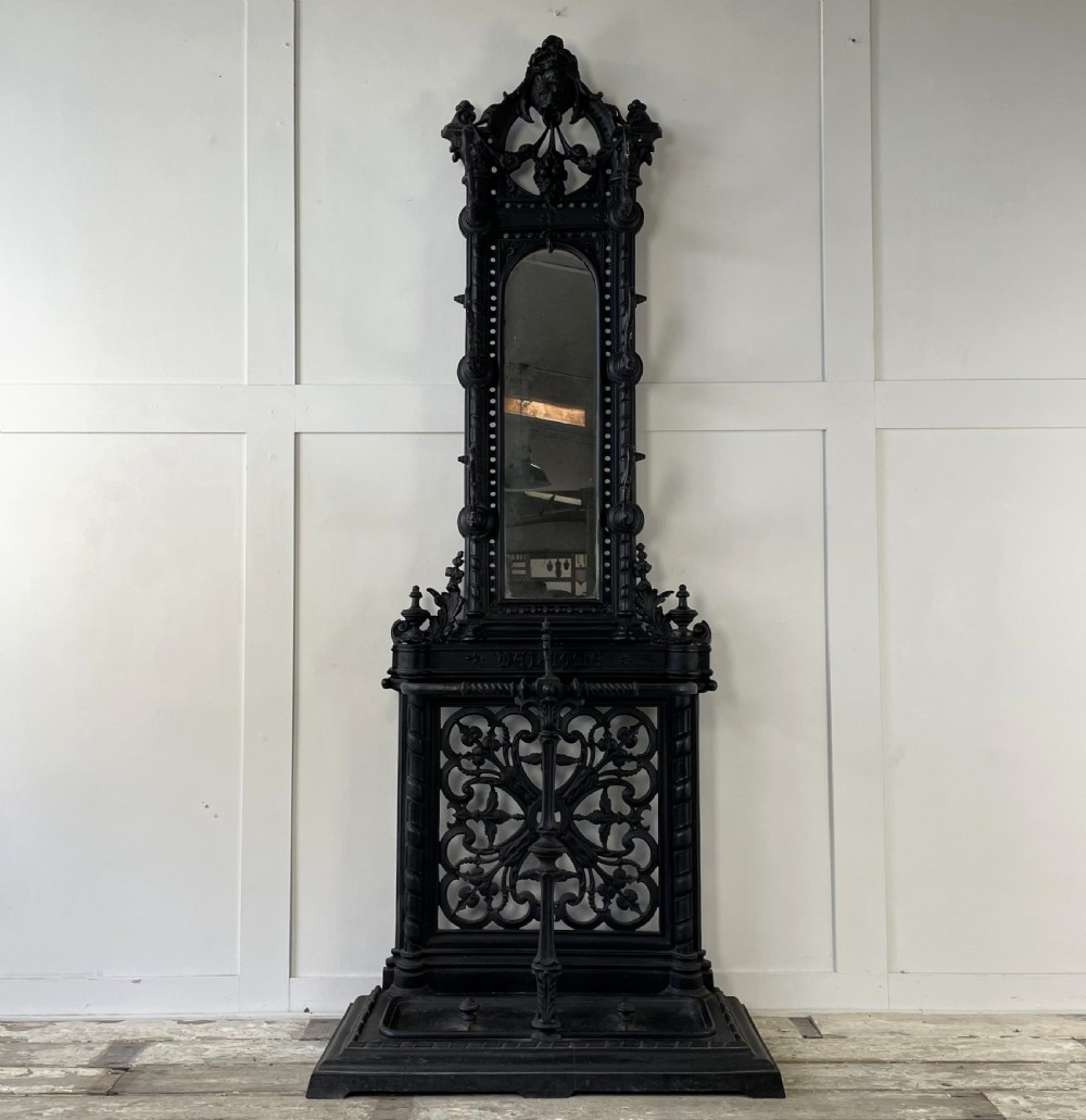 A tall black cast iron Victorian hall stand featuring an integrated central mirror, branching coat hooks, scrollwork tracery, and an umbrella drip tray at the base. View II