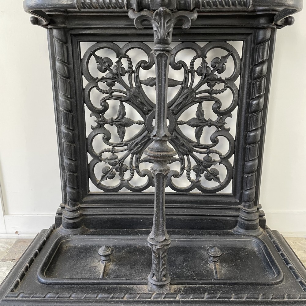 A tall black cast iron Victorian hall stand featuring an integrated central mirror, branching coat hooks, scrollwork tracery, and an umbrella drip tray at the base. View XIV