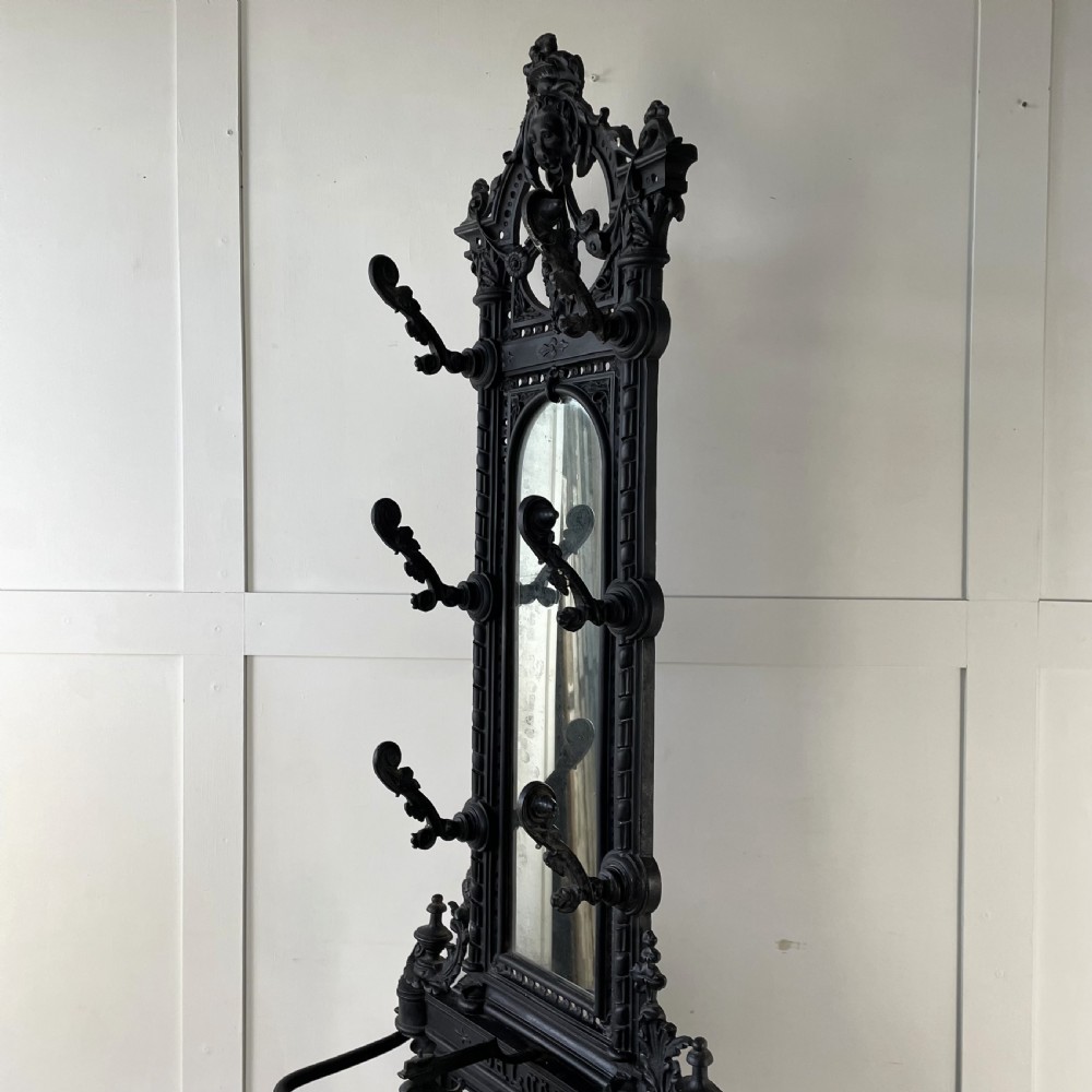 A tall black cast iron Victorian hall stand featuring an integrated central mirror, branching coat hooks, scrollwork tracery, and an umbrella drip tray at the base. View XVII