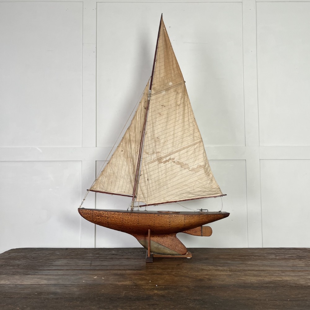 An Edwardian wooden pond yacht with a carved timber hull, tall rigged mast, cream fabric sails, and a wooden stand.
