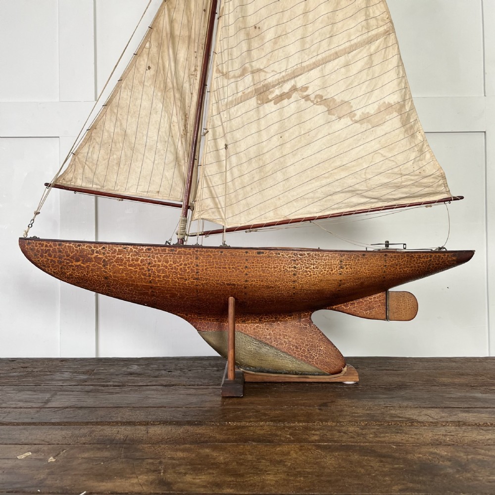 An Edwardian wooden pond yacht with a carved timber hull, tall rigged mast, cream fabric sails, and a wooden stand. View II