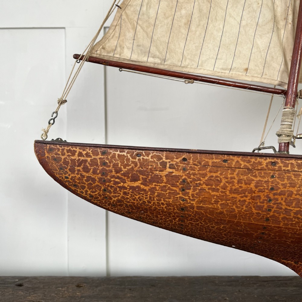 An Edwardian wooden pond yacht with a carved timber hull, tall rigged mast, cream fabric sails, and a wooden stand. View III