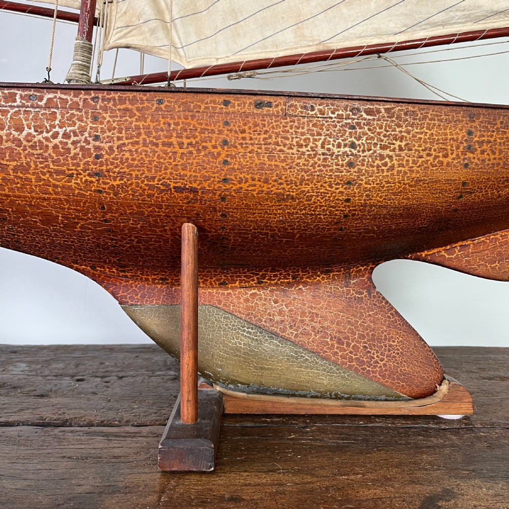 An Edwardian wooden pond yacht with a carved timber hull, tall rigged mast, cream fabric sails, and a wooden stand. View IV