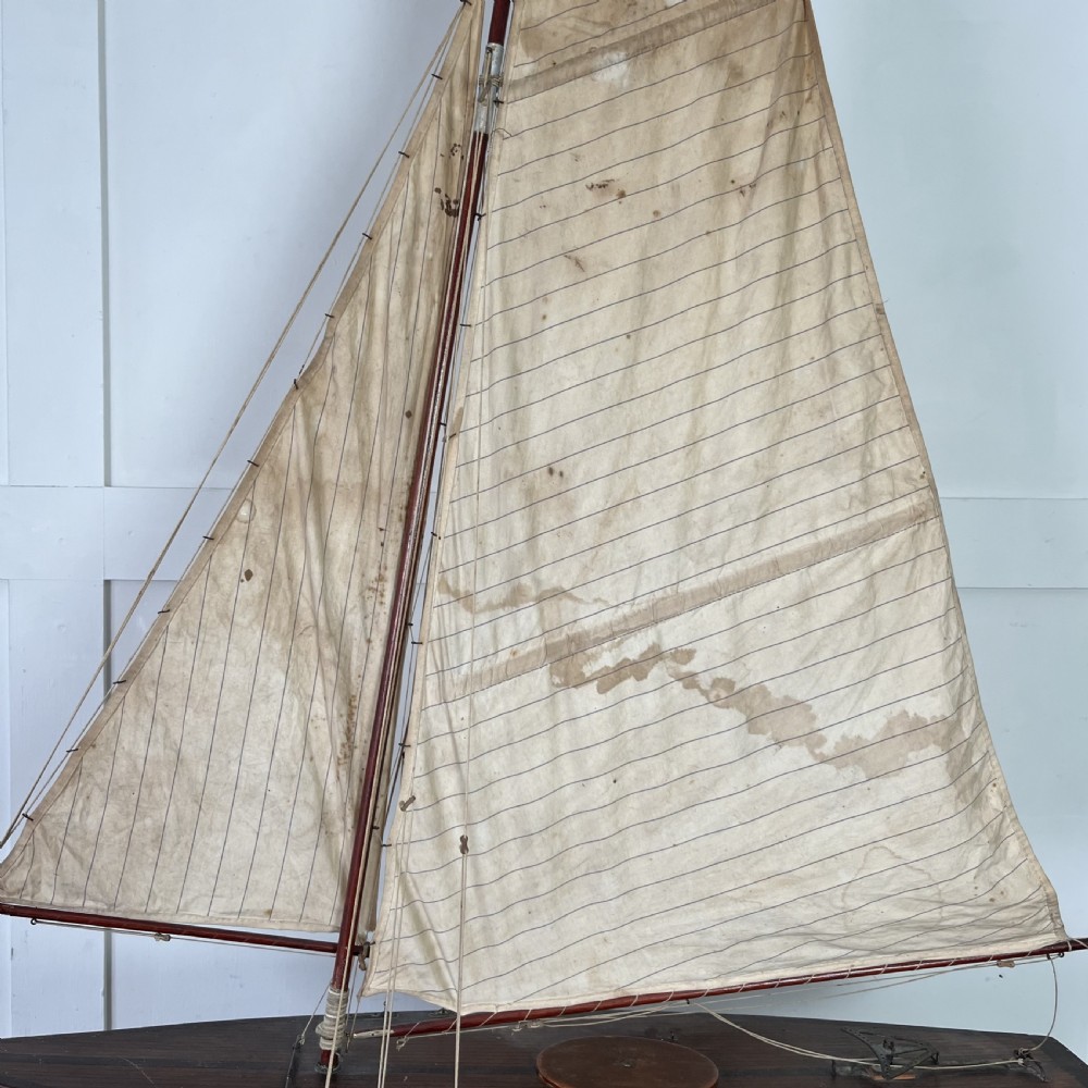 An Edwardian wooden pond yacht with a carved timber hull, tall rigged mast, cream fabric sails, and a wooden stand. View IX