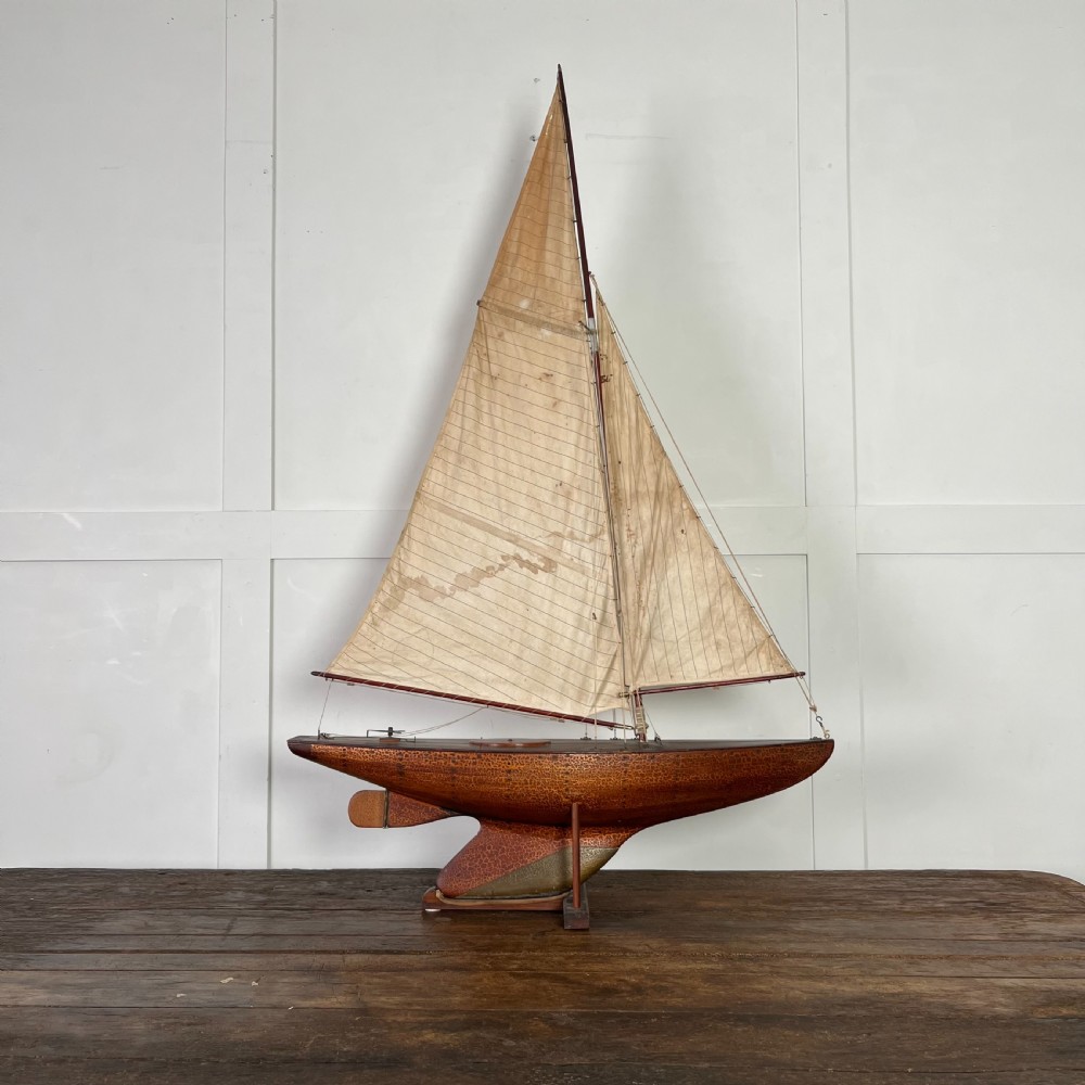 An Edwardian wooden pond yacht with a carved timber hull, tall rigged mast, cream fabric sails, and a wooden stand. View X