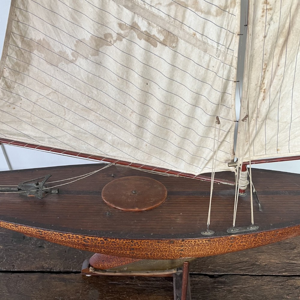 An Edwardian wooden pond yacht with a carved timber hull, tall rigged mast, cream fabric sails, and a wooden stand. View XV