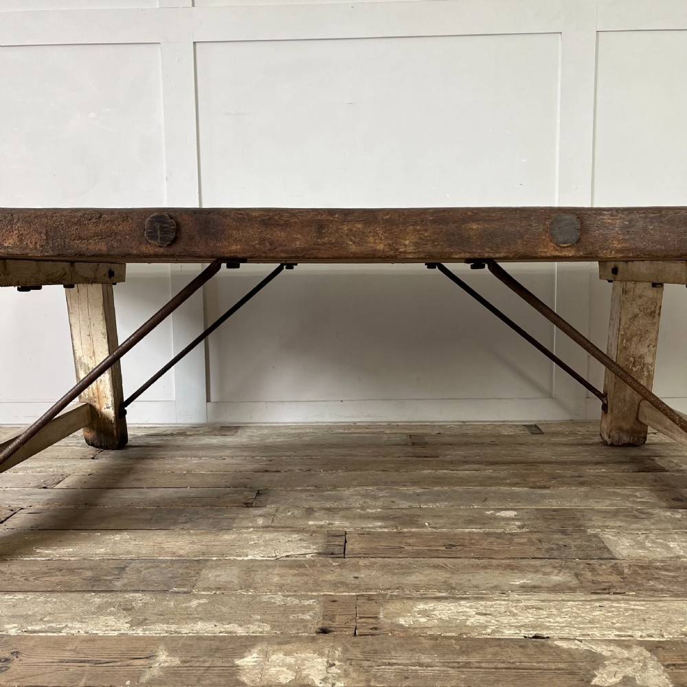 An early 20th-century rustic cheese press table featuring a thick dark wooden top and distressed painted trestle legs with metal bracing. View X