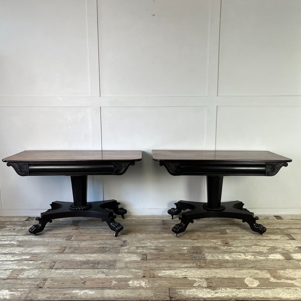 A pair of William IV ebonised mahogany console tables with polished mahogany tops, central pedestal columns, and carved claw feet on brass castors.