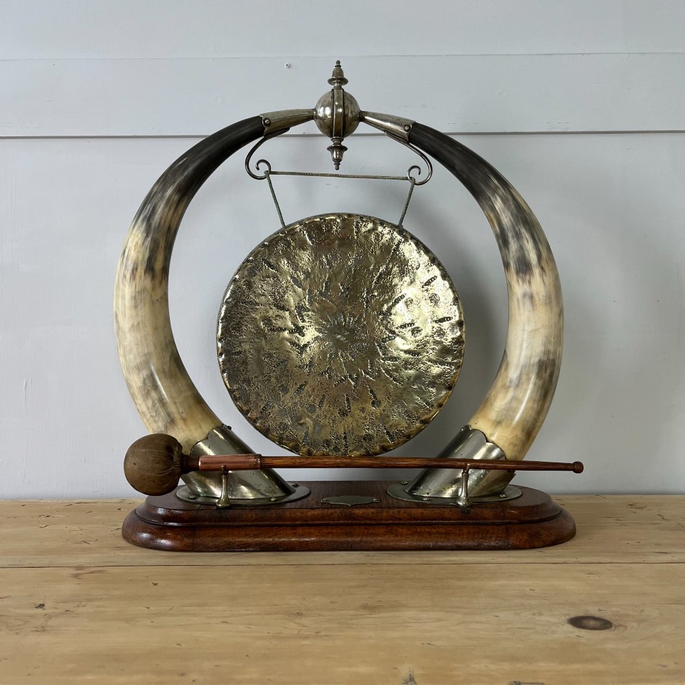 A Victorian tabletop dinner gong featuring two curved natural horns mounted on a polished oak base, holding a central hammered brass disc with an accompanying wooden striker.
