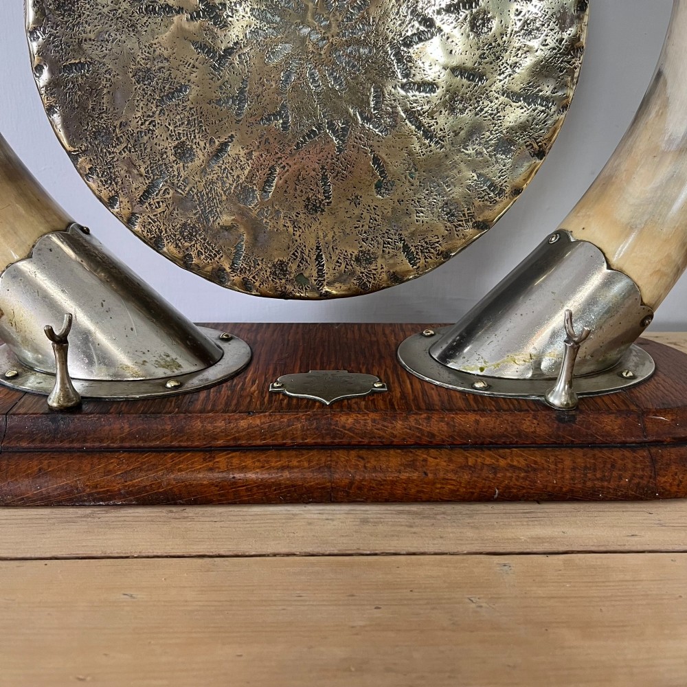 A Victorian tabletop dinner gong featuring two curved natural horns mounted on a polished oak base, holding a central hammered brass disc with an accompanying wooden striker. View IV