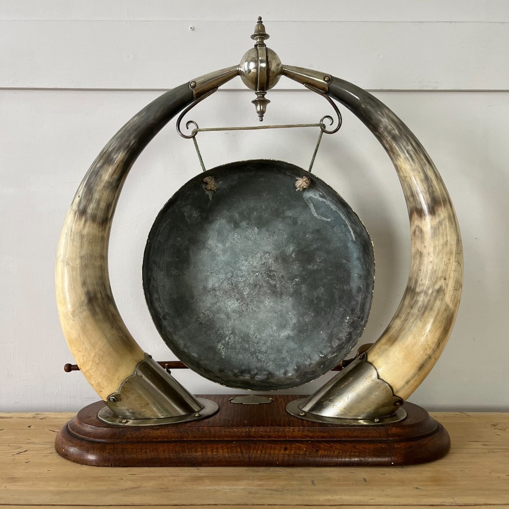 A Victorian tabletop dinner gong featuring two curved natural horns mounted on a polished oak base, holding a central hammered brass disc with an accompanying wooden striker. View VII