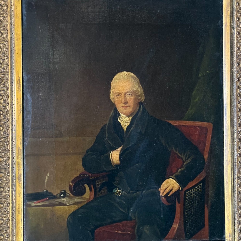 A 19th-century oil portrait depicting a seated gentleman in dark attire against a dark background, set within a heavily carved giltwood frame. View II