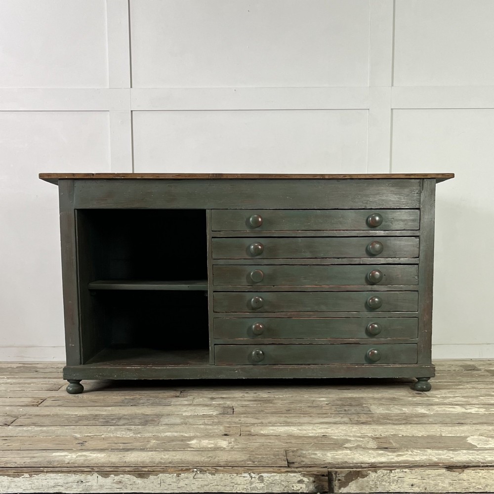 A low Victorian dark green painted pine artist chest featuring six shallow drawers on the right, an open shelf compartment on the left, and an unpainted timber top.