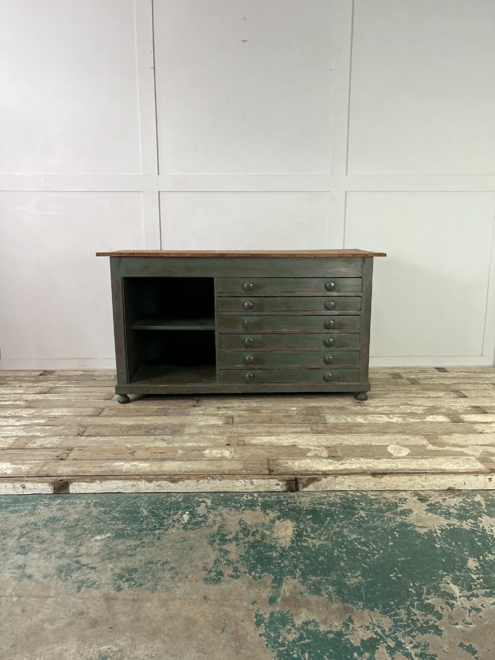 A low Victorian dark green painted pine artist chest featuring six shallow drawers on the right, an open shelf compartment on the left, and an unpainted timber top. View II