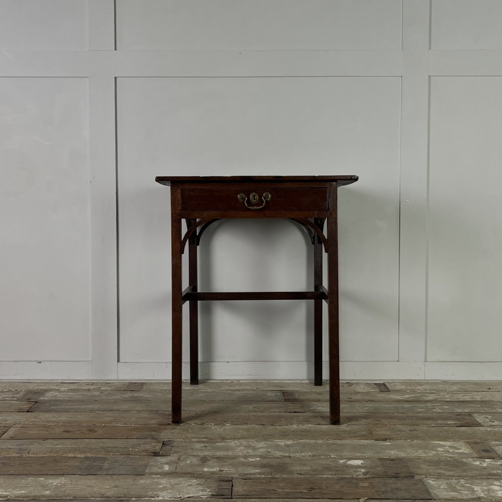 An early 19th century dark oak side table featuring a single drawer with a brass handle, arched aprons, and straight square legs connected by a low stretcher.