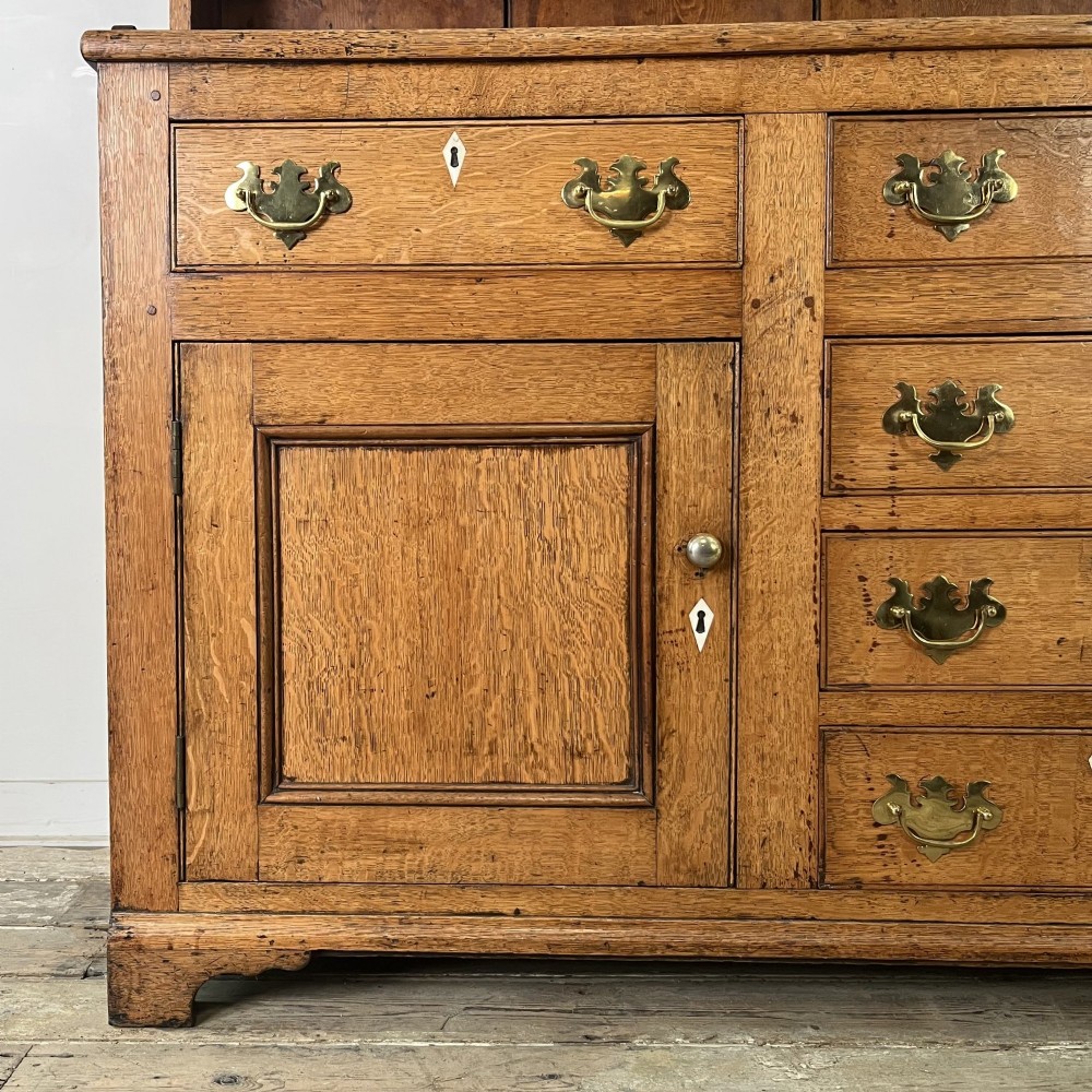 An early nineteenth-century oak kitchen dresser featuring an open upper plate rack over a base with central drawers and side cupboard doors with brass hardware. View V