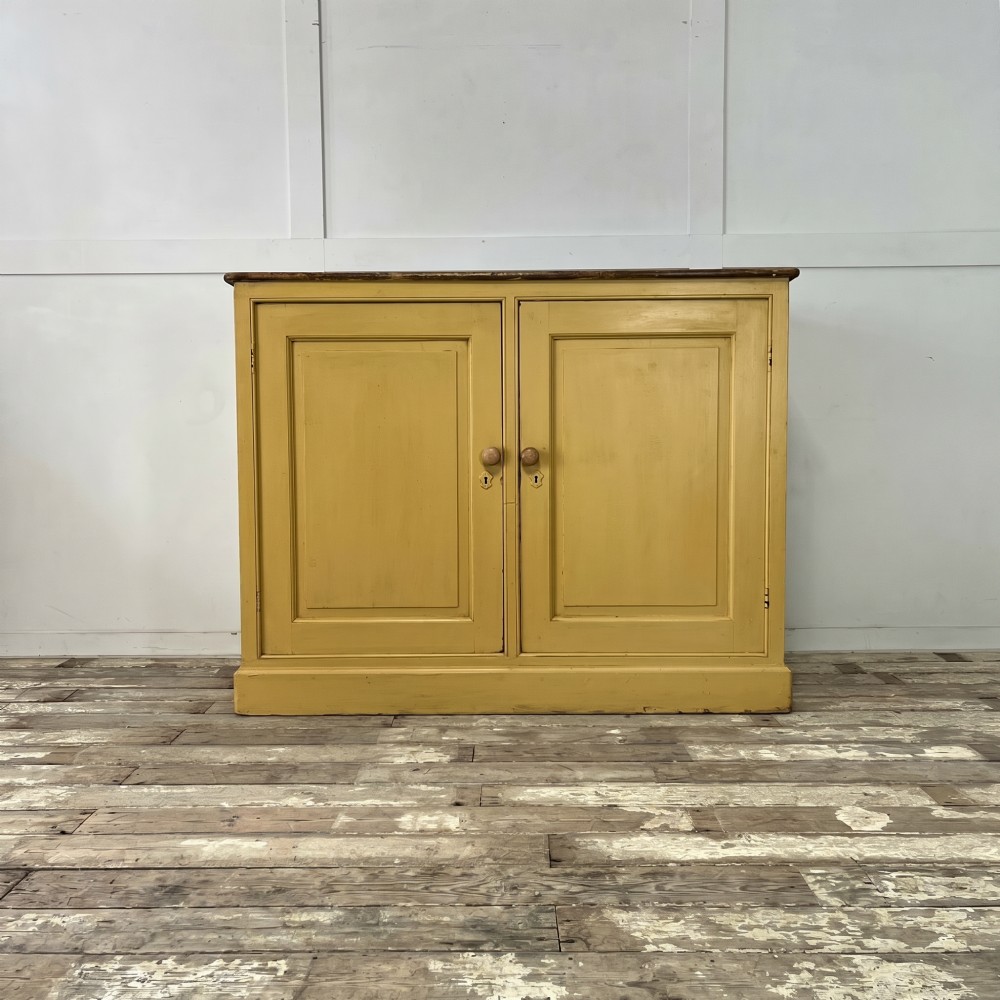 A large Victorian pine school cupboard featuring a mustard yellow painted finish, two panelled doors with wooden knobs, and a dark waxed wooden top.