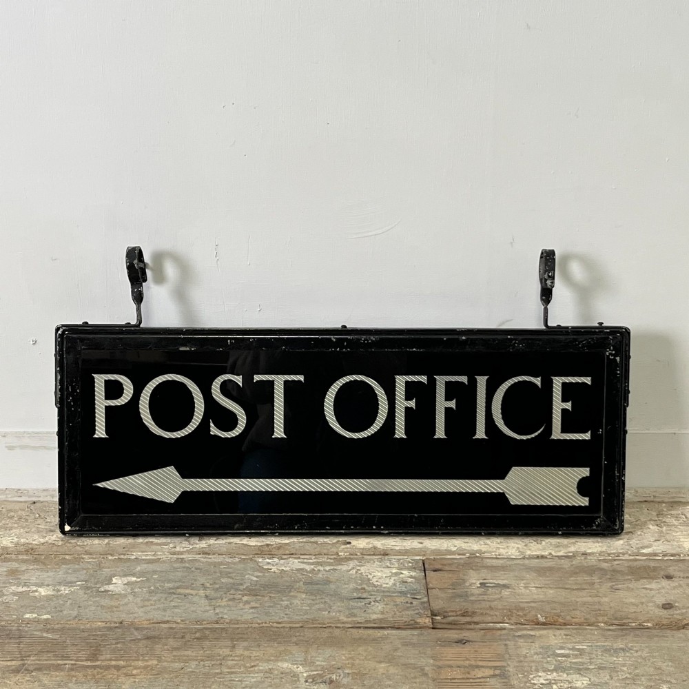A rectangular double-sided black glass directional sign featuring white lettering that reads POST OFFICE above a striped directional arrow, mounted in a dark iron frame with top loops.