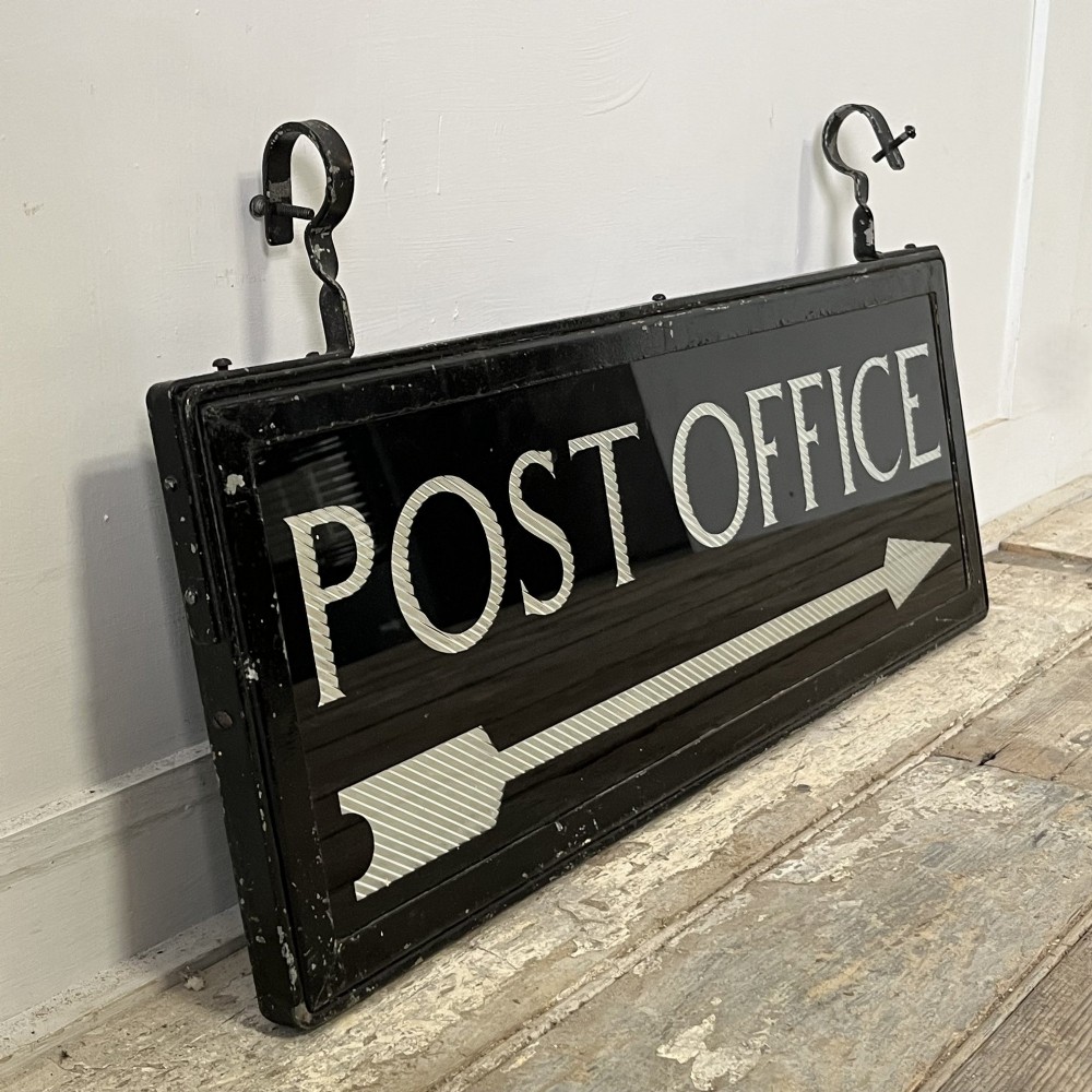A rectangular double-sided black glass directional sign featuring white lettering that reads POST OFFICE above a striped directional arrow, mounted in a dark iron frame with top loops. View V