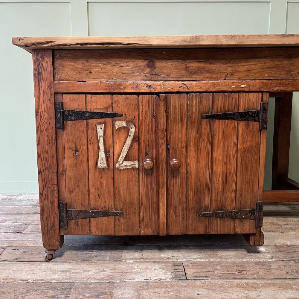 An early twentieth-century rustic pine bakers prep table with dual cupboard doors, black iron strap hinges, painted number twelve, and wooden castors. View V