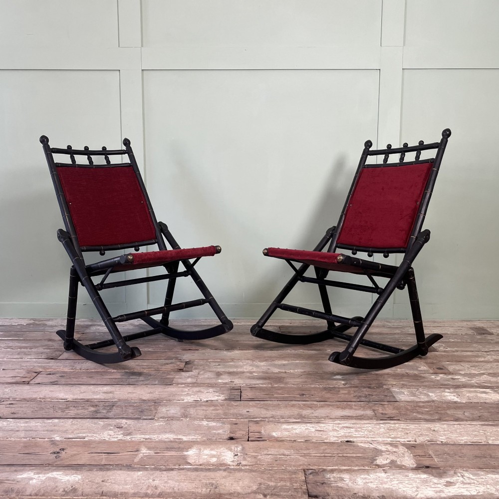 A pair of Victorian ebonised wood folding rocking chairs featuring faux bamboo turned frames and deep red velvet upholstered seats and backrests.