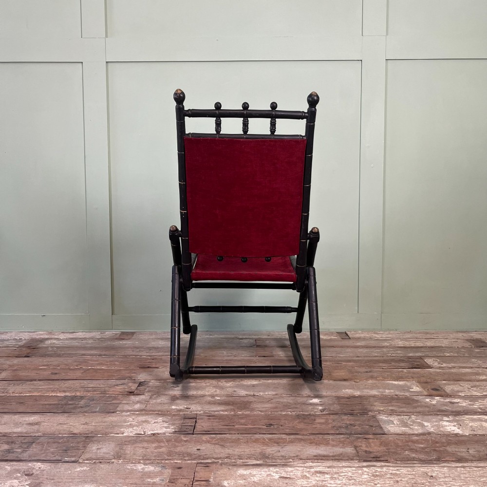 A pair of Victorian ebonised wood folding rocking chairs featuring faux bamboo turned frames and deep red velvet upholstered seats and backrests. View VII