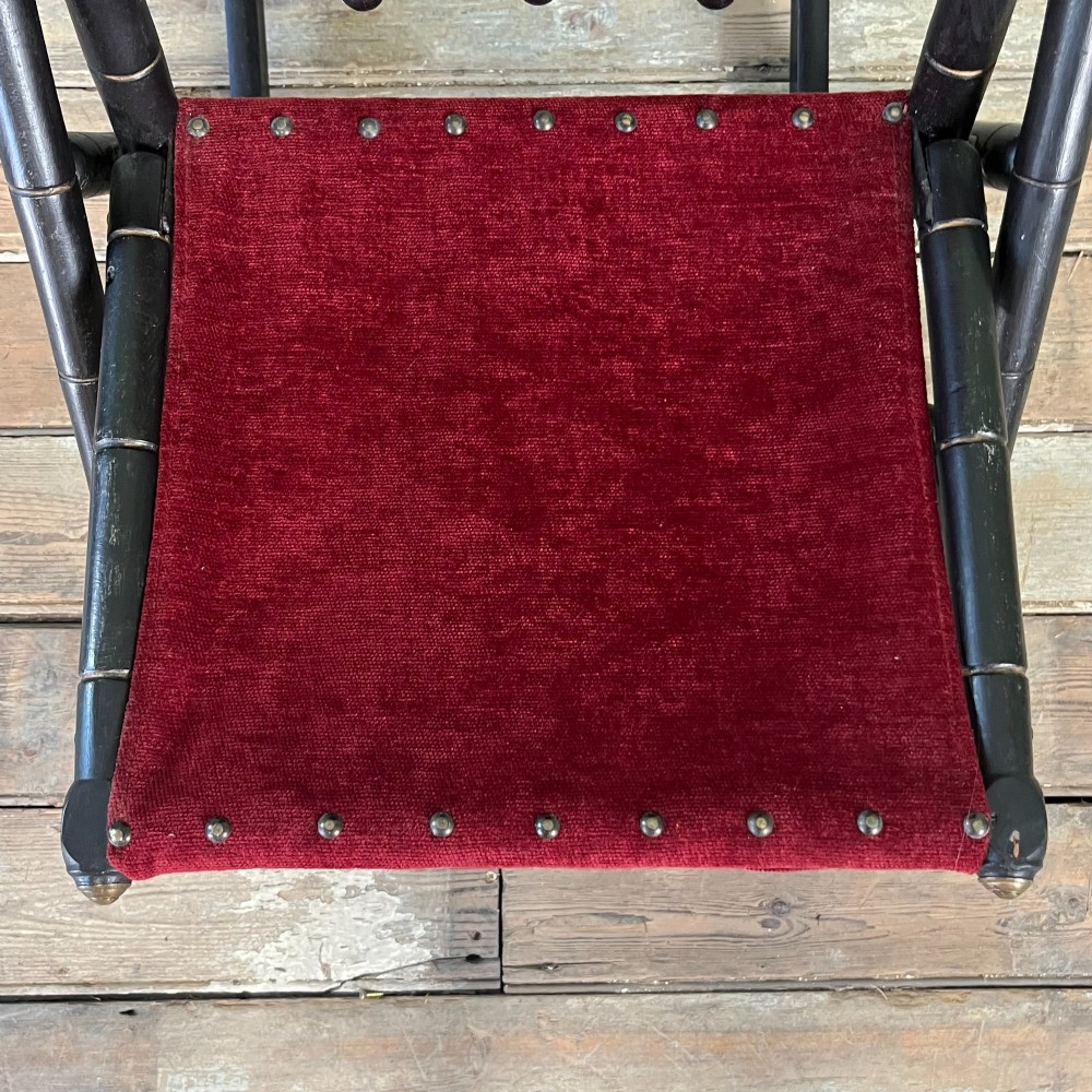 A pair of Victorian ebonised wood folding rocking chairs featuring faux bamboo turned frames and deep red velvet upholstered seats and backrests. View X