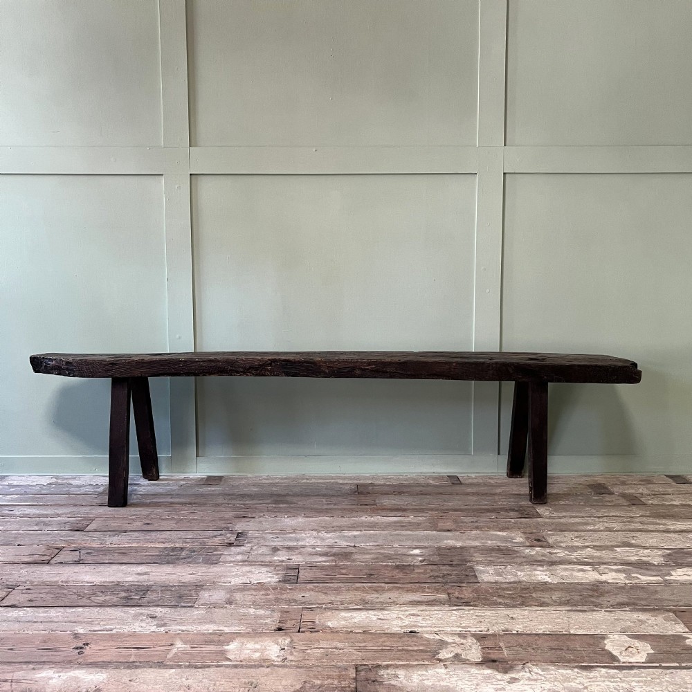 A long rustic dark oak bench featuring an unrefined thick slab seat supported by four simple splayed legs.