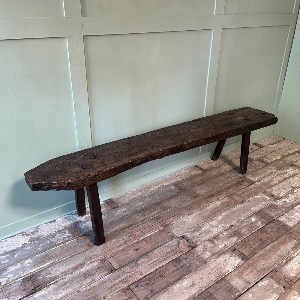 A long rustic dark oak bench featuring an unrefined thick slab seat supported by four simple splayed legs. View II