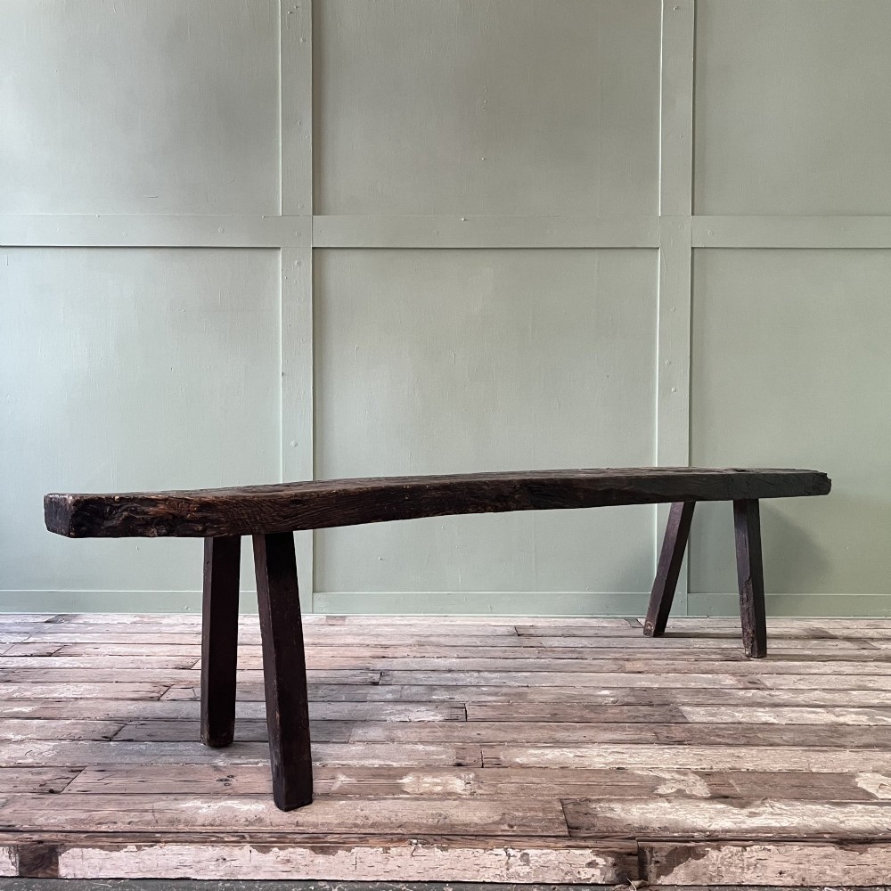 A long rustic dark oak bench featuring an unrefined thick slab seat supported by four simple splayed legs. View X