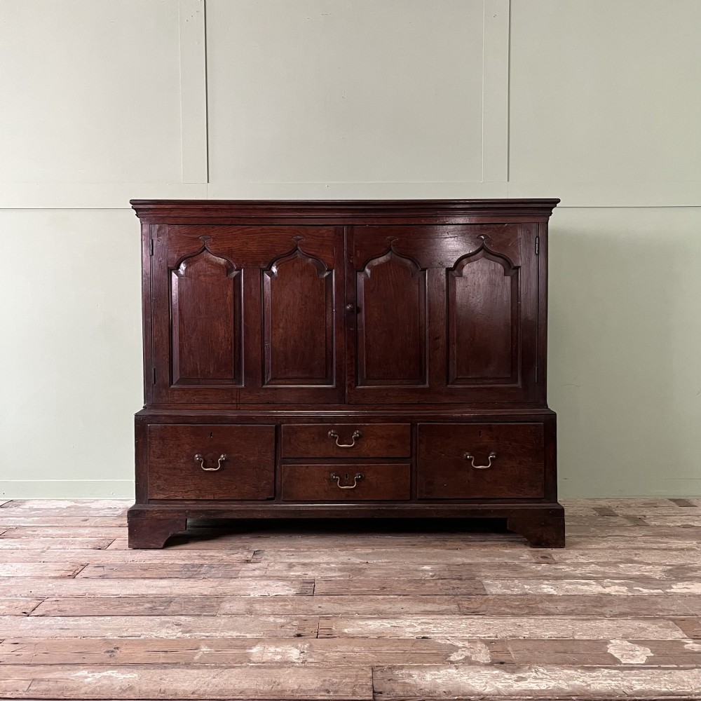 An 18th-century dark oak low linen press cupboard featuring arched fielded panel doors over a base with four drawers and brass handles.
