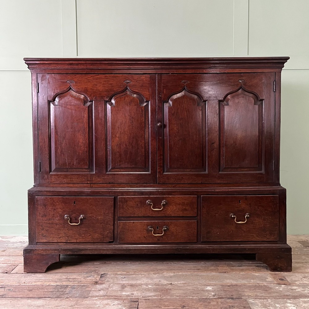 An 18th-century dark oak low linen press cupboard featuring arched fielded panel doors over a base with four drawers and brass handles. View II