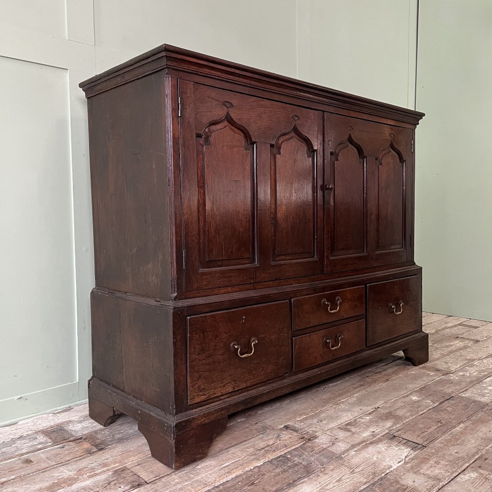 An 18th-century dark oak low linen press cupboard featuring arched fielded panel doors over a base with four drawers and brass handles. View III