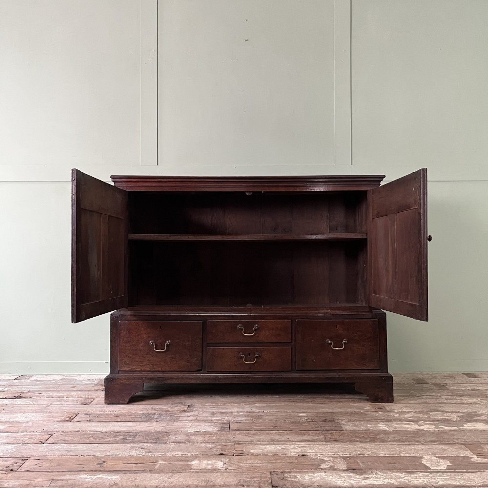 An 18th-century dark oak low linen press cupboard featuring arched fielded panel doors over a base with four drawers and brass handles. View IV