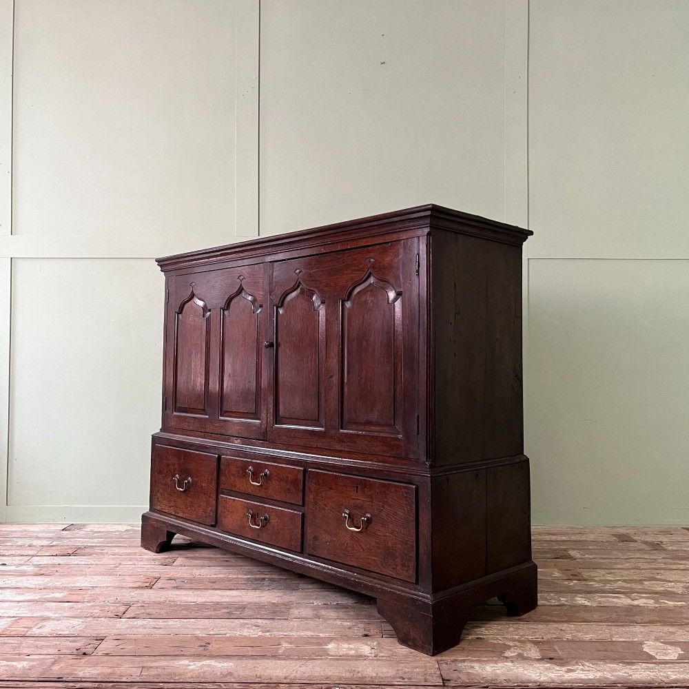 An 18th-century dark oak low linen press cupboard featuring arched fielded panel doors over a base with four drawers and brass handles. View V