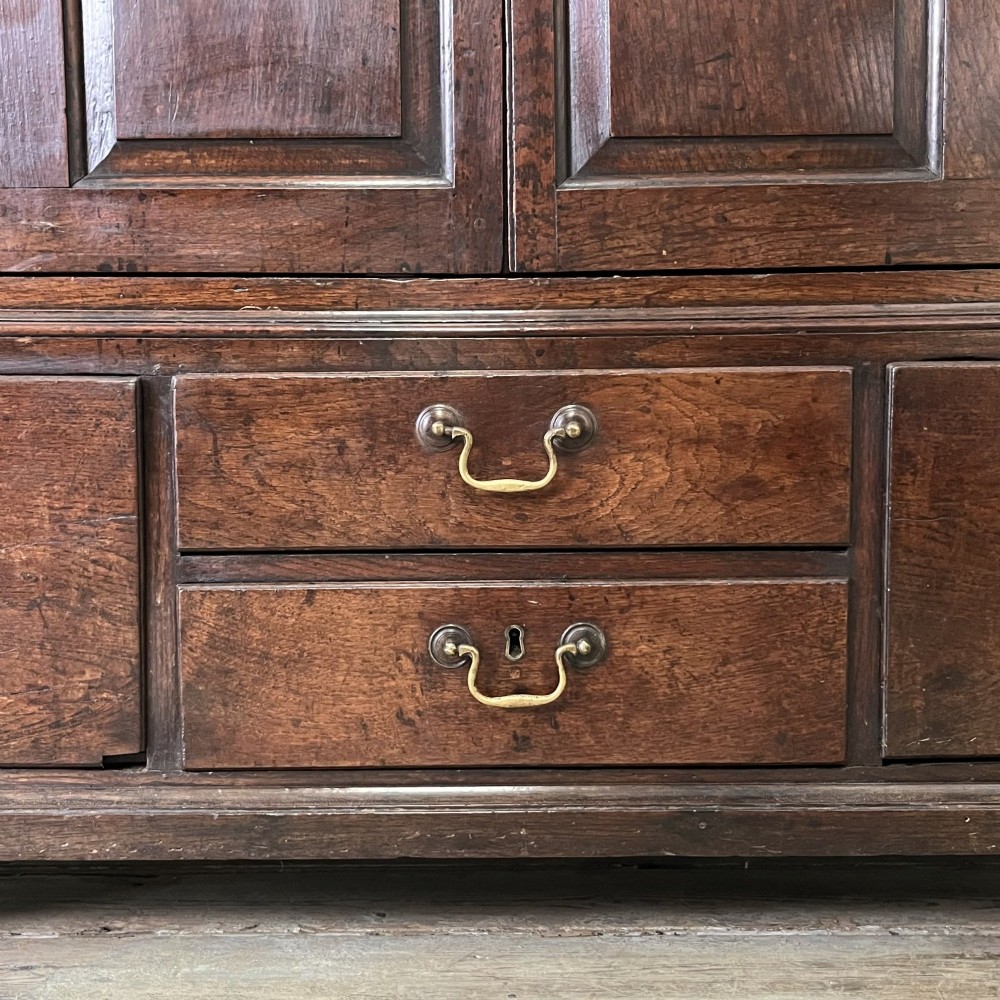 An 18th-century dark oak low linen press cupboard featuring arched fielded panel doors over a base with four drawers and brass handles. View XV