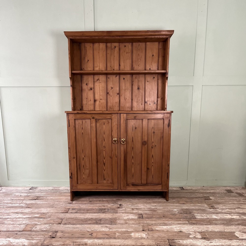 An early twentieth-century pine kitchen dresser featuring an open top display rack with two shelves above a two-door cabinet base with brass ring handles.